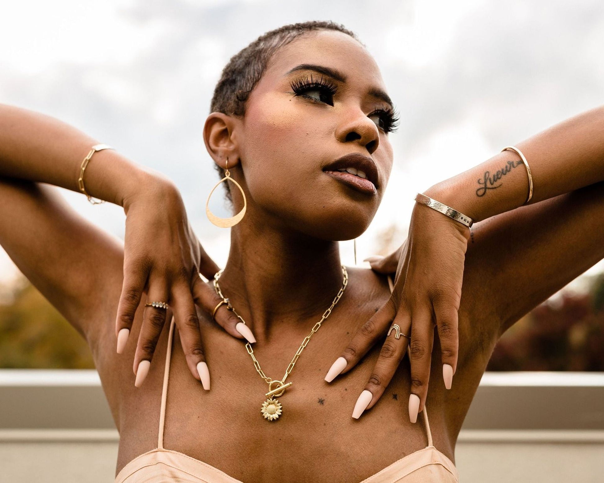 A woman with short hair and dark skin poses with her hands on her chest, wearing a gold necklace and earrings.