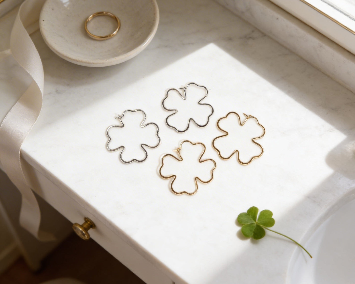 a white marble countertop with a pair of gold earrings shaped like four-leaf clovers, a small white bowl, and a single green clover leaf.