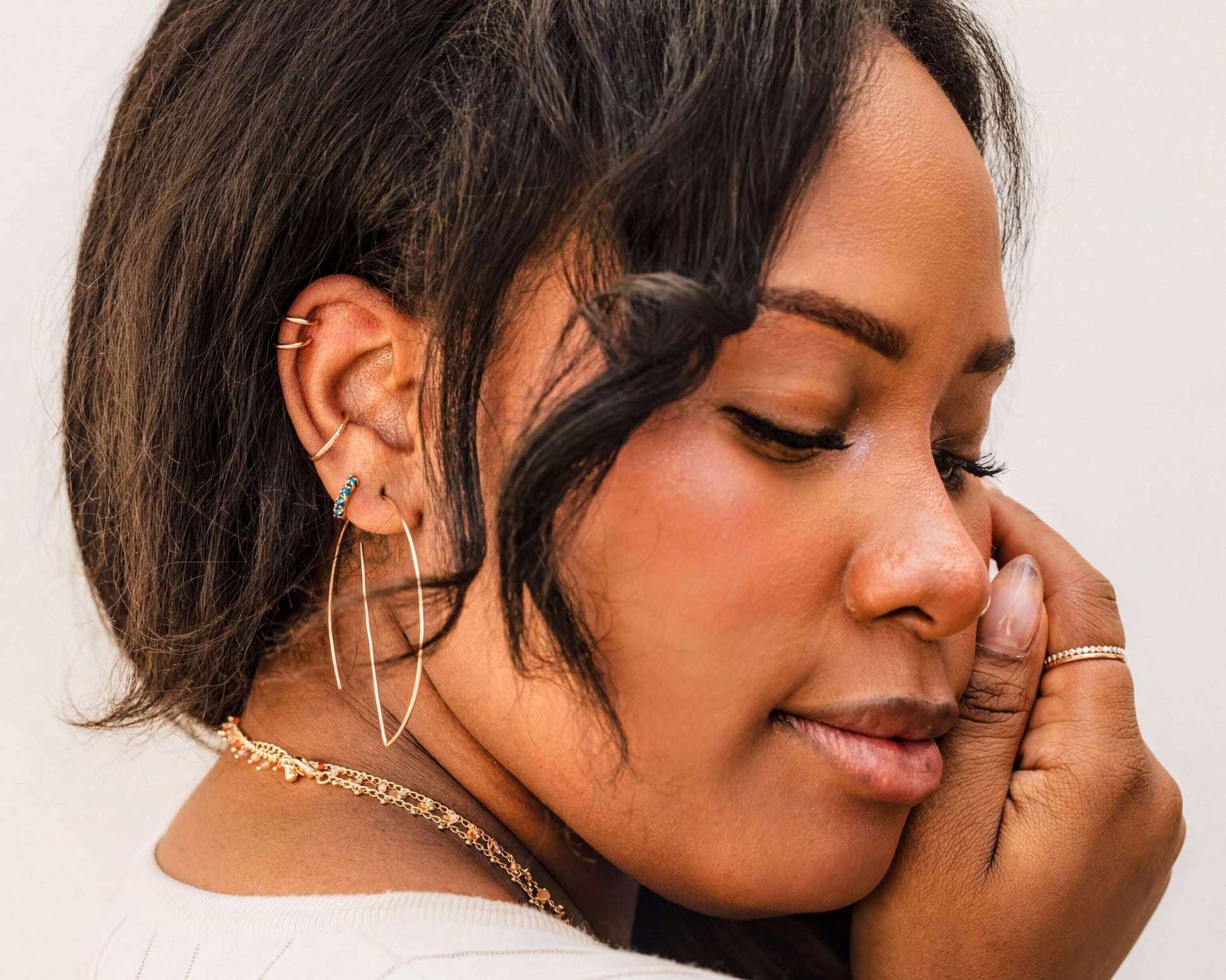 A close-up portrait of a woman with dark hair, wearing multiple earrings and jewelry, including a large hoop earring and a ring on her finger.