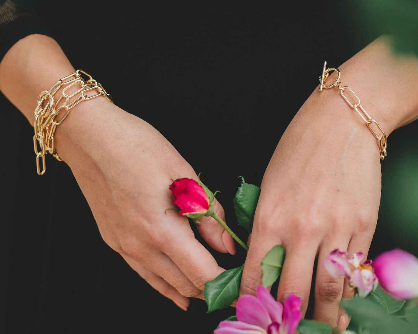 A close-up image of a person's hands wearing gold bracelets and holding a red rose, with some pink flowers in the background.