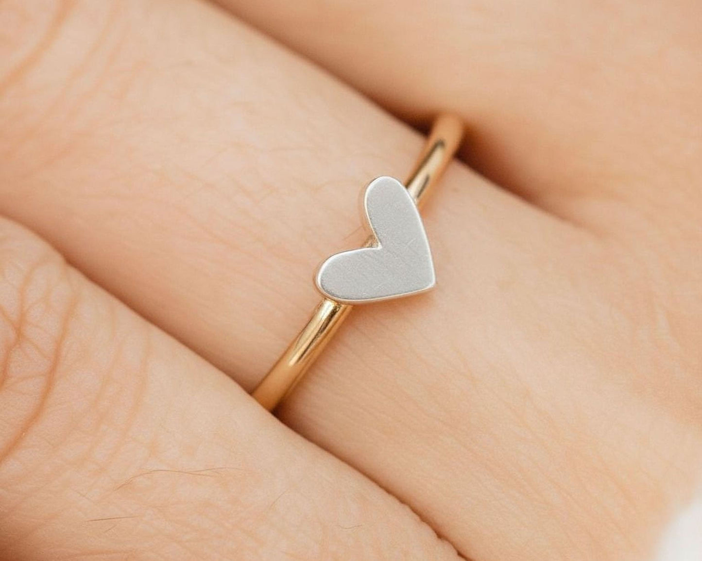 A close-up image of a person's hand wearing a gold ring with a small silver heart-shaped charm.
