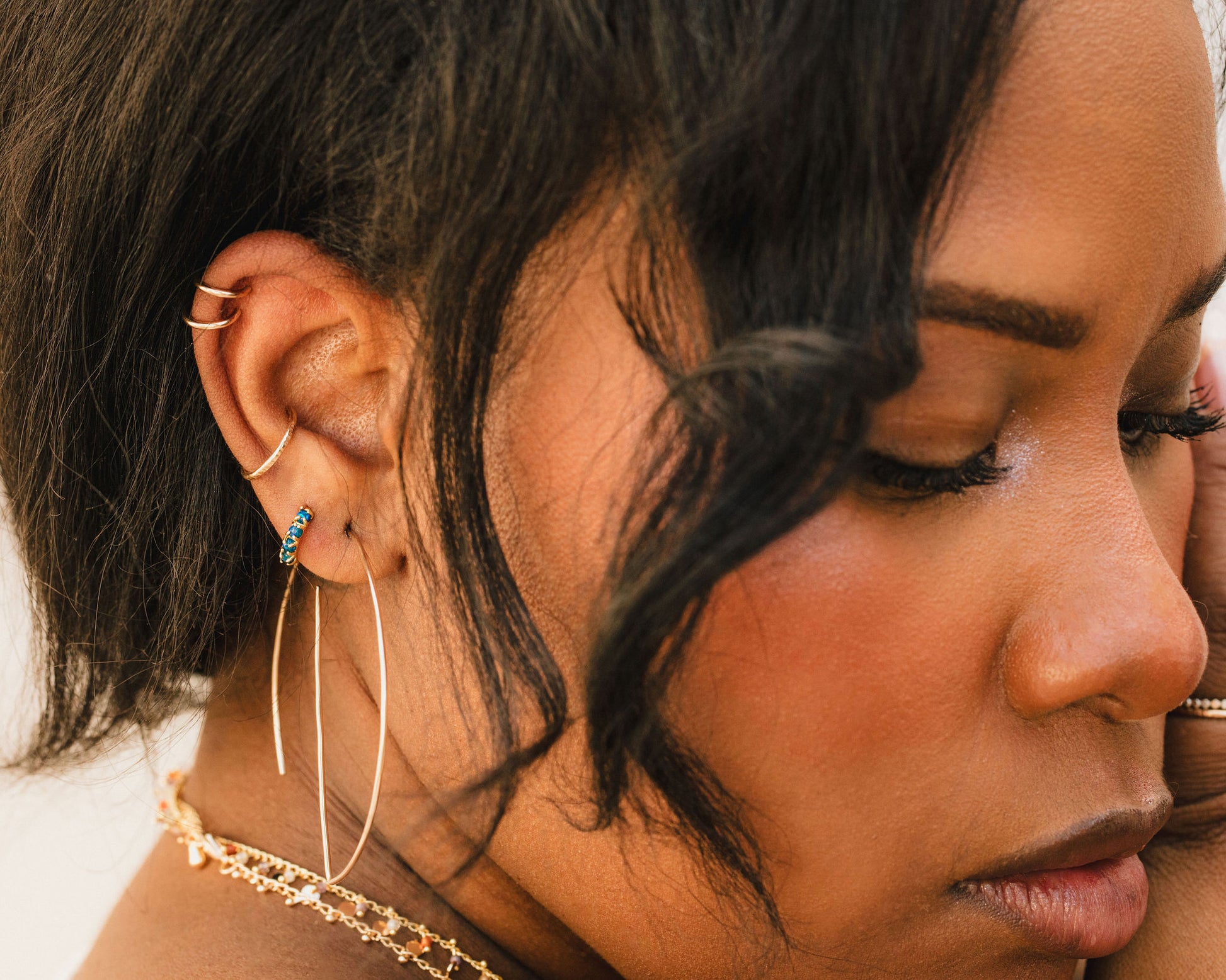 A close-up of a woman's face, with her eyes closed and her hair styled in loose waves. She is wearing large hoop earrings and a necklace.