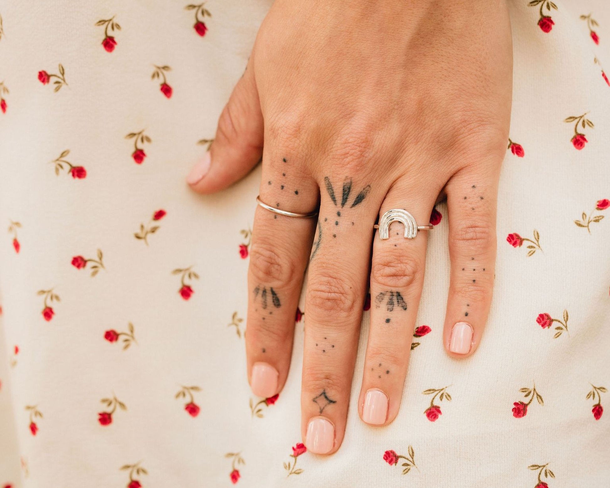 A close-up of a person's hand with multiple tattoos on the fingers, wearing rings, and resting on a white fabric with a red floral pattern.