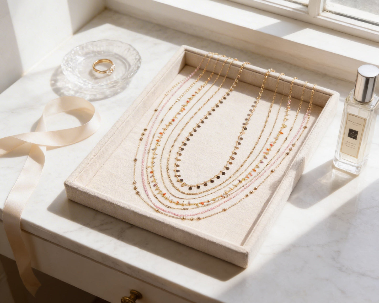 A beige jewelry tray holds a collection of necklaces, with a clear glass plate holding a ring and a bottle of perfume placed next to it.