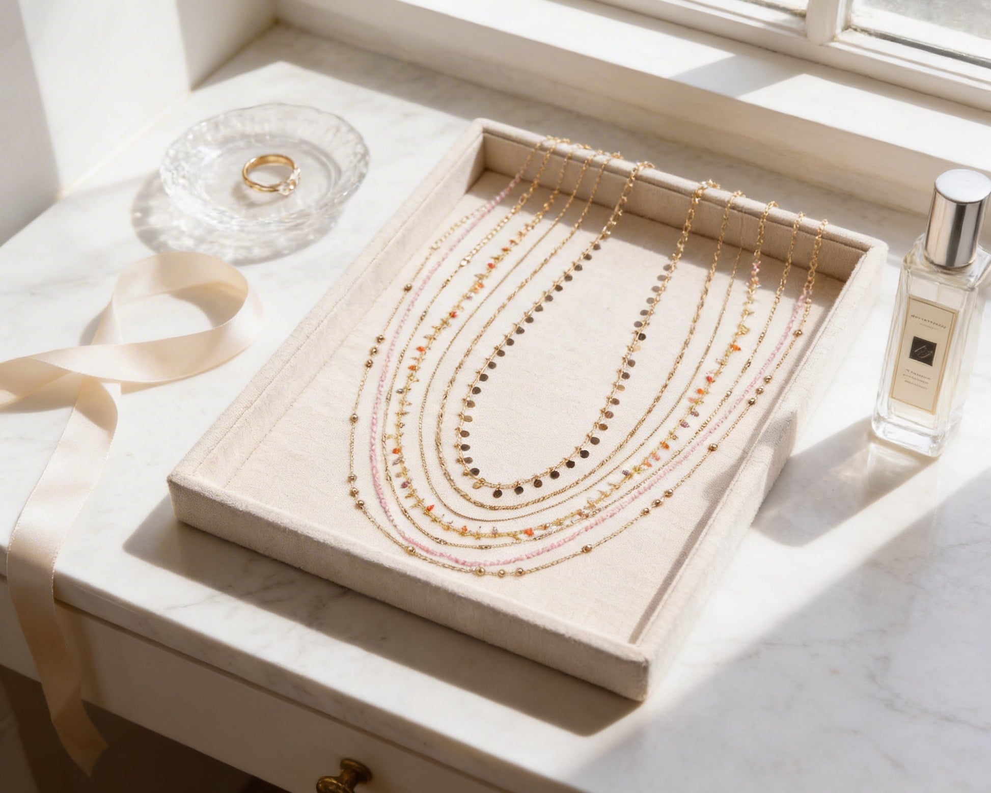 A beige jewelry tray holds a collection of necklaces, with a clear glass plate holding a ring and a bottle of perfume placed next to it.