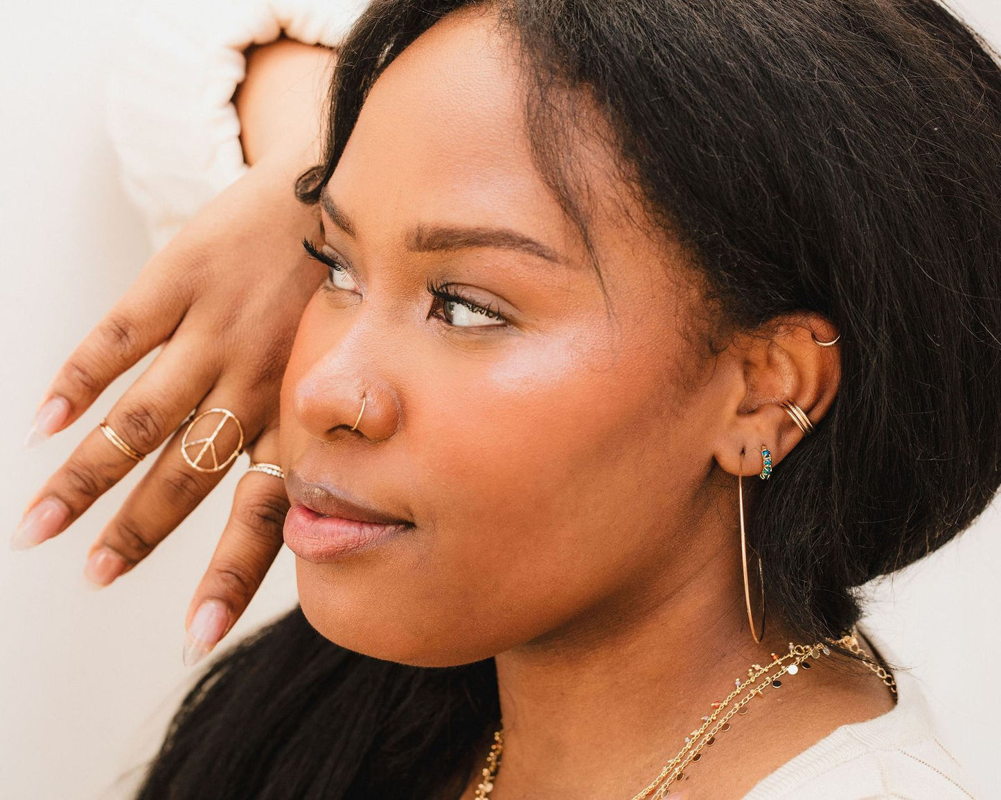 A close-up portrait of a woman with dark hair, wearing multiple rings and earrings, and looking off to the side with a thoughtful expression.