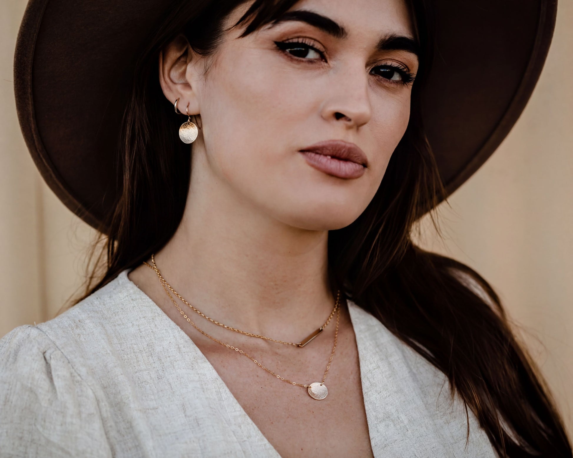 A woman wearing a large brown hat and a necklace with multiple pendants, posing for a portrait.