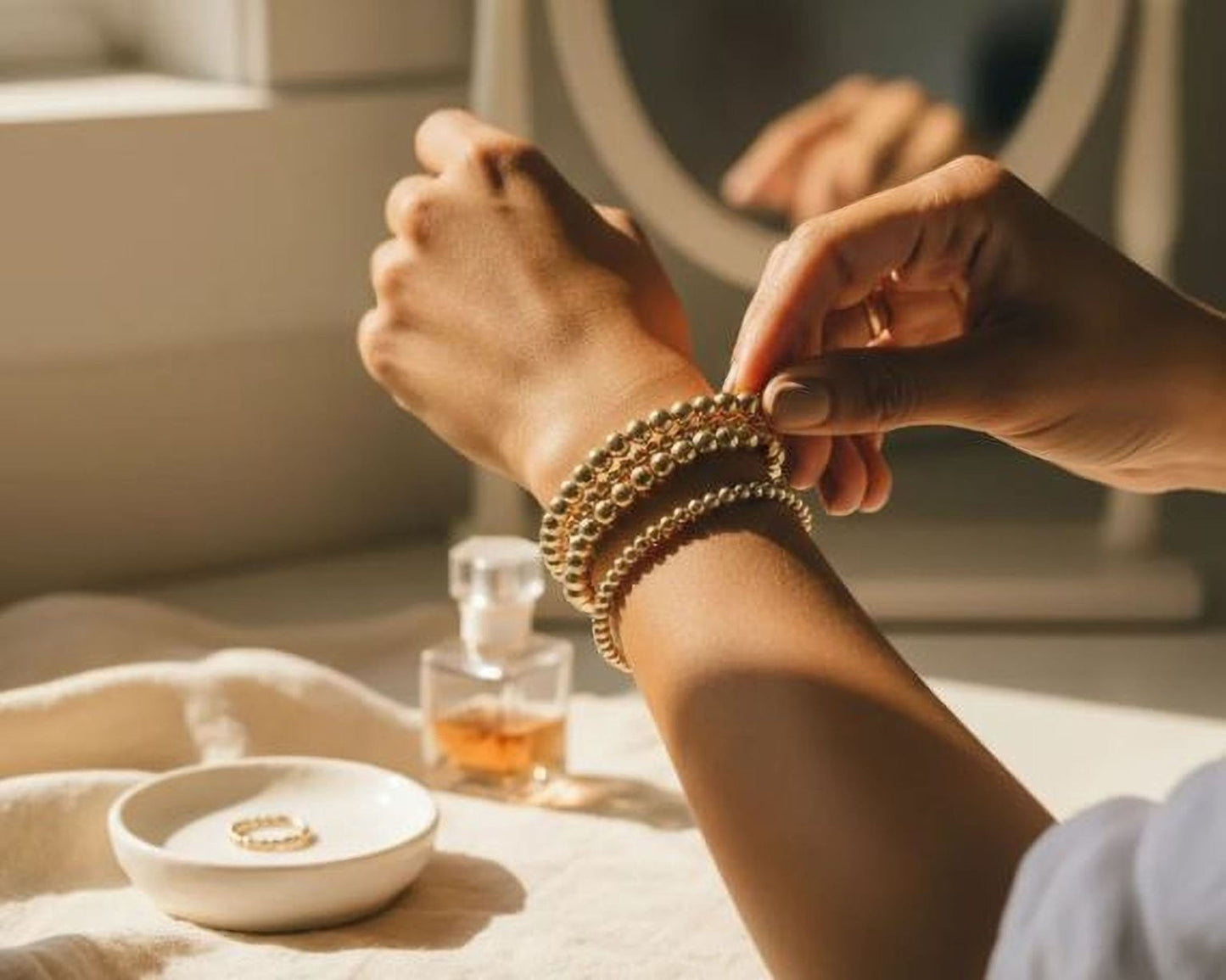 A person's hands are adjusting a bracelet on their wrist, with a mirror and a small bowl of jewelry visible in the background.
