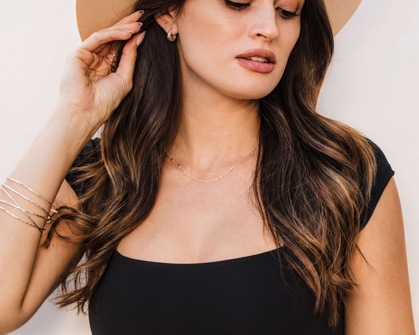 A woman with long, wavy brown hair wearing a black top and a wide-brimmed hat, with her hand touching her ear.