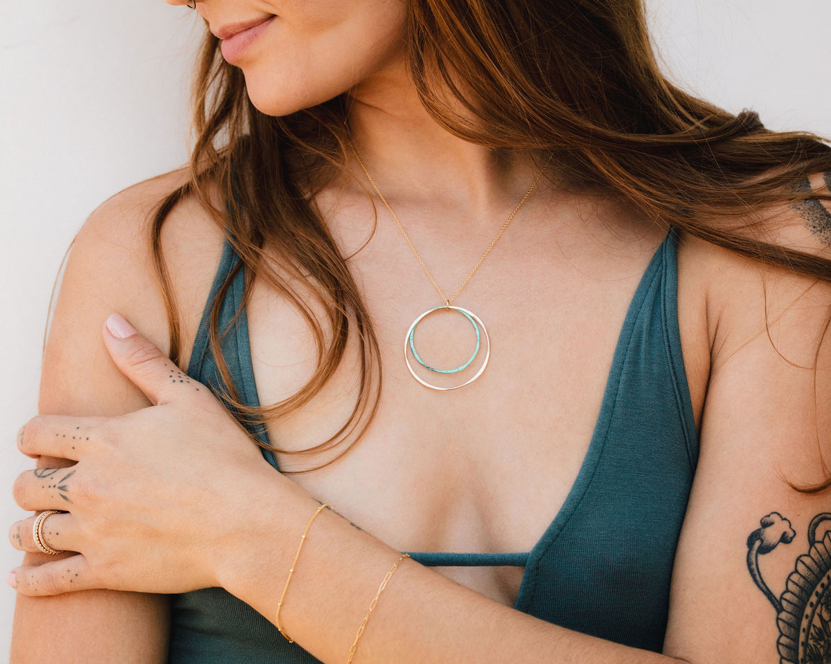 A woman with long brown hair wearing a green top and a necklace with a circular pendant stands with her arms crossed.