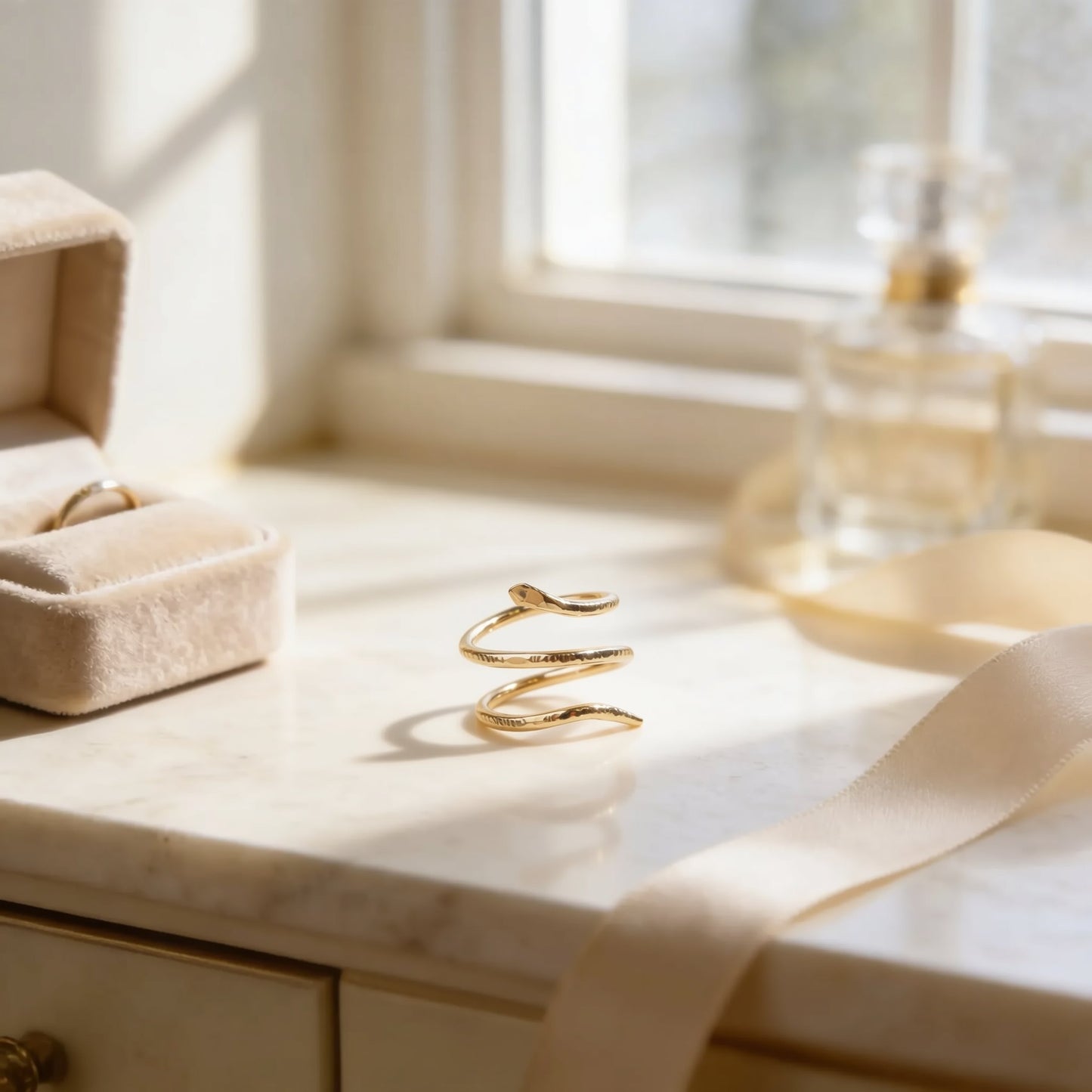 a gold ring, a gold necklace, and a bottle of perfume on a marble countertop, with a window in the background.