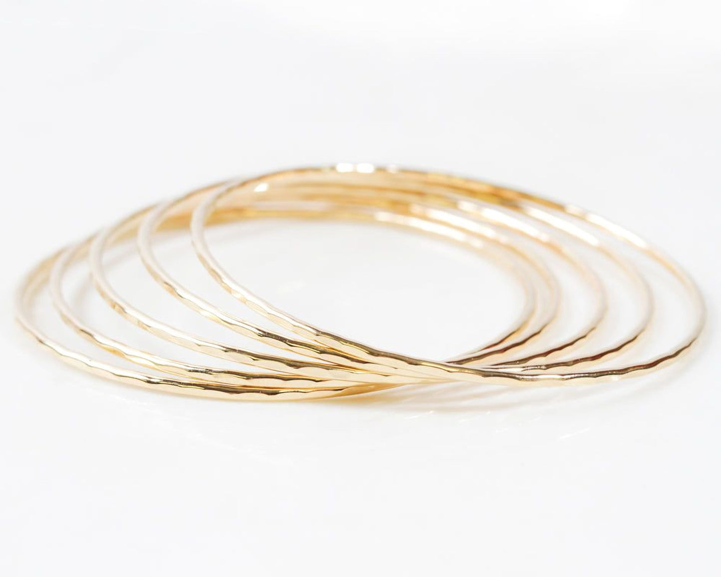 Image shows close up of the exquisite texture of our 14 karat yellow gold filled bangles. Showing five stunning bangles, each bracelet is hand hammered which gives them their special sparkle. Wear individually or stack them up to create a unique set.