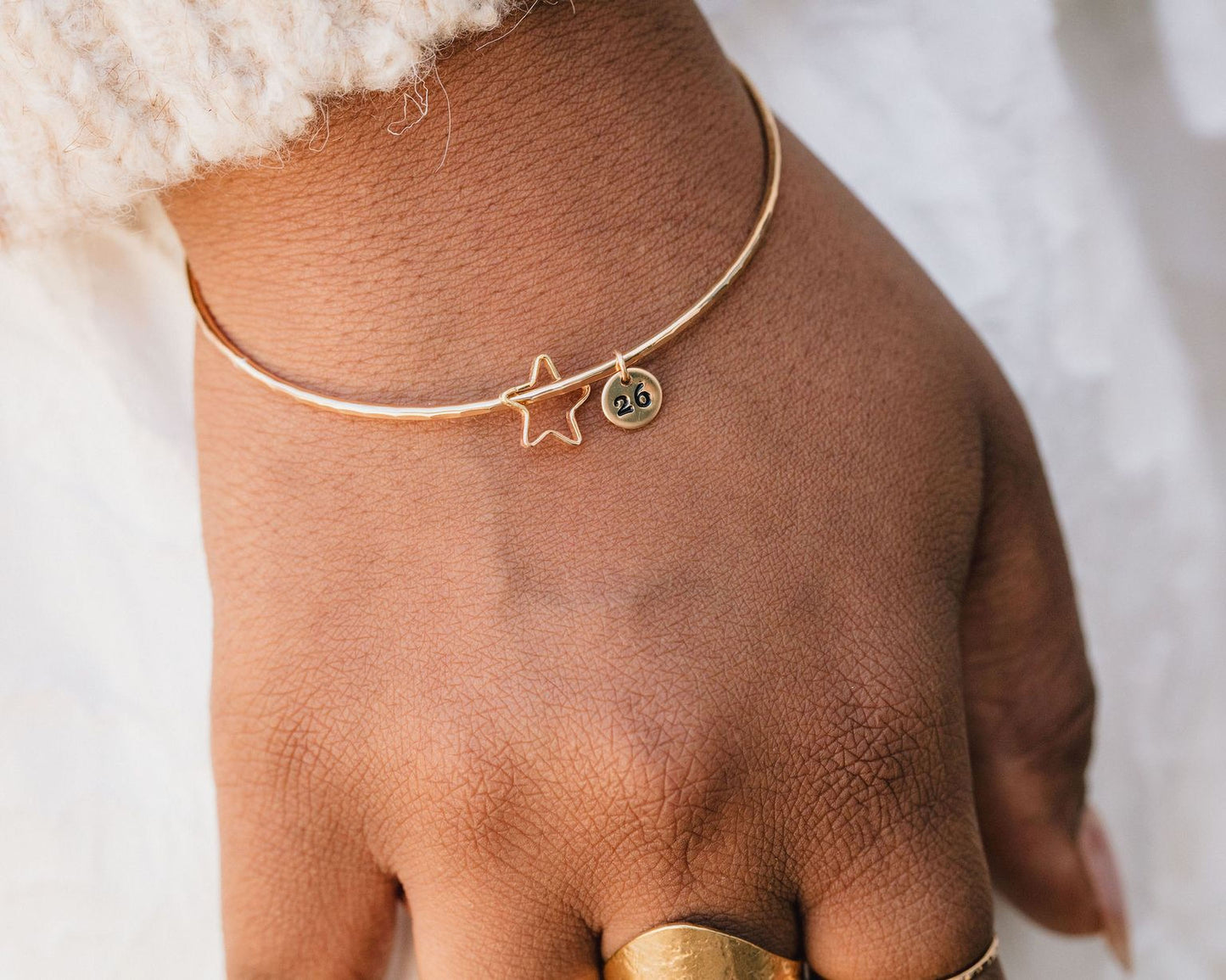 A close-up view of a person's hand wearing a gold bangle bracelet with a star charm and the number "26" engraved on it.