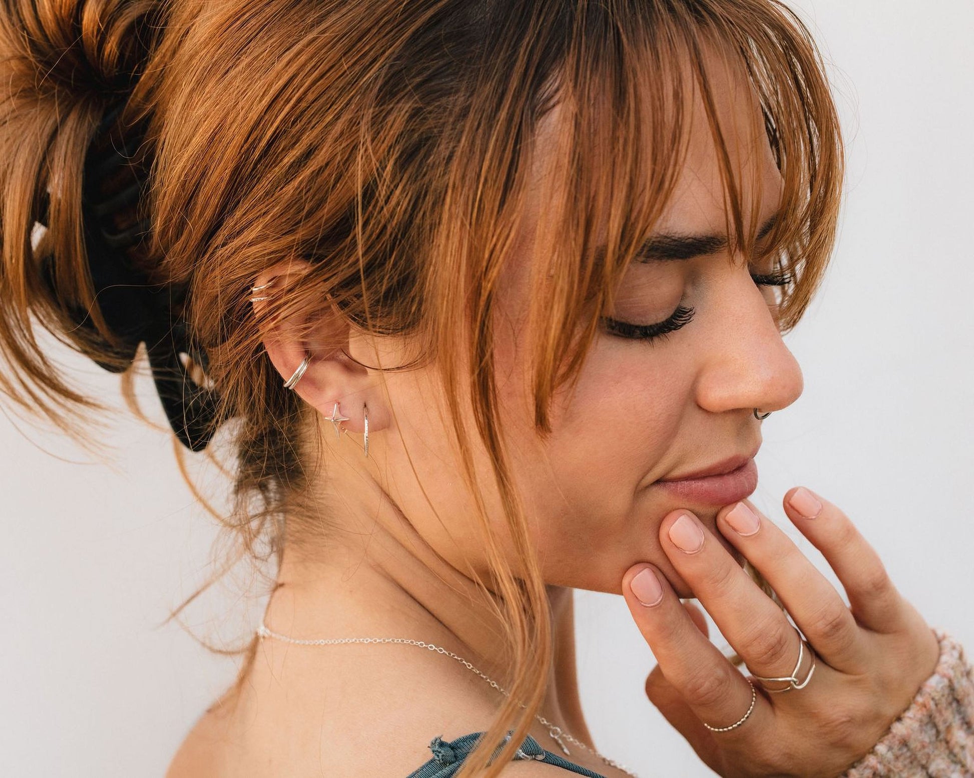 A woman with long, reddish-brown hair is shown in profile, her eyes closed and her hand resting on her chin. She is wearing multiple earrings and a nose ring, and her hair is styled in a messy bun.