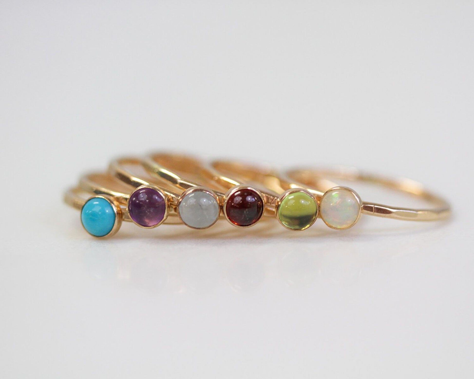 A row of gold rings with colorful gemstones, including turquoise, amethyst, garnet, and opal, arranged on a white surface.