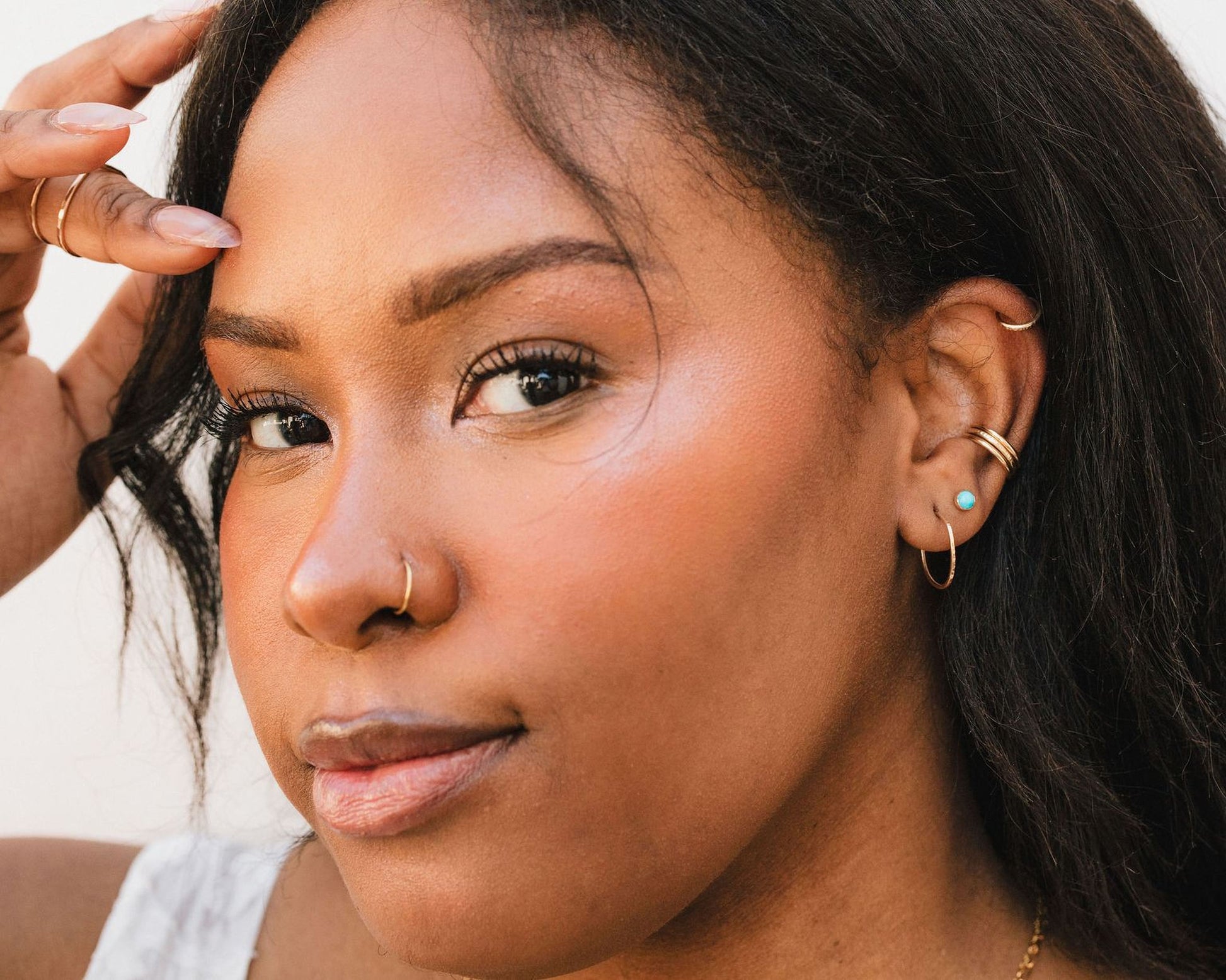 A close-up portrait of a woman with dark hair, wearing multiple earrings and a nose ring.