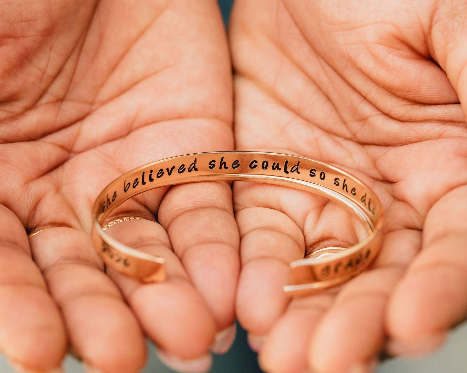 A person's hands holding a gold-colored bracelet with the text "she believed she could so she did" engraved on it.