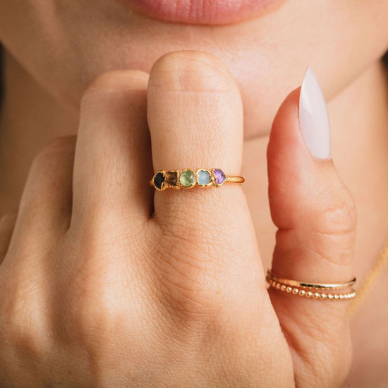 A close-up of a person's hand wearing a gold ring with colorful gemstones, and another ring on the same finger.