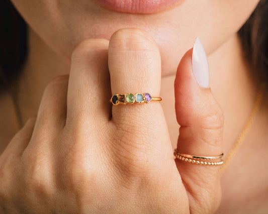 A close-up of a person's hand wearing a gold ring with colorful gemstones, and another ring on the same finger.
