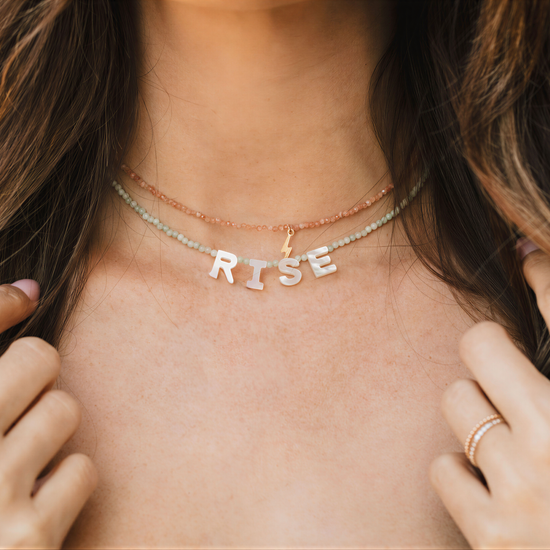 A close-up of a person's neck and chest area, showing a necklace with the word "RISE" spelled out in letters.