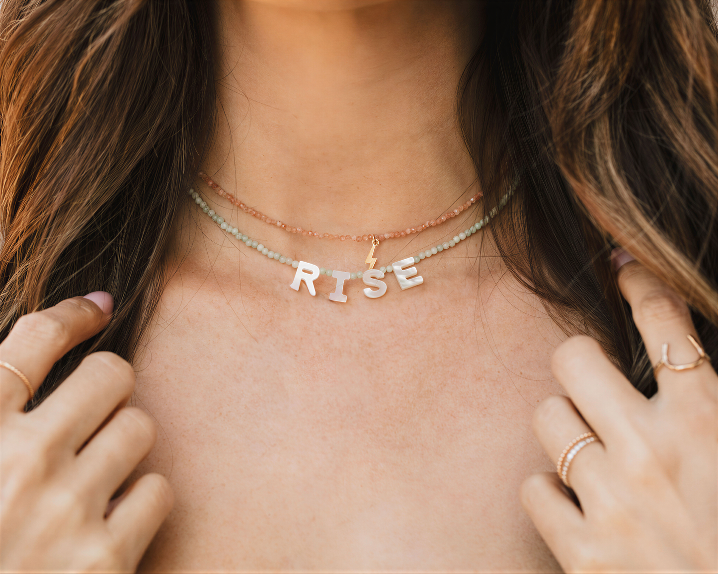 A close-up of a person's neck and chest area, showing a necklace with the word "RISE" spelled out in letters.