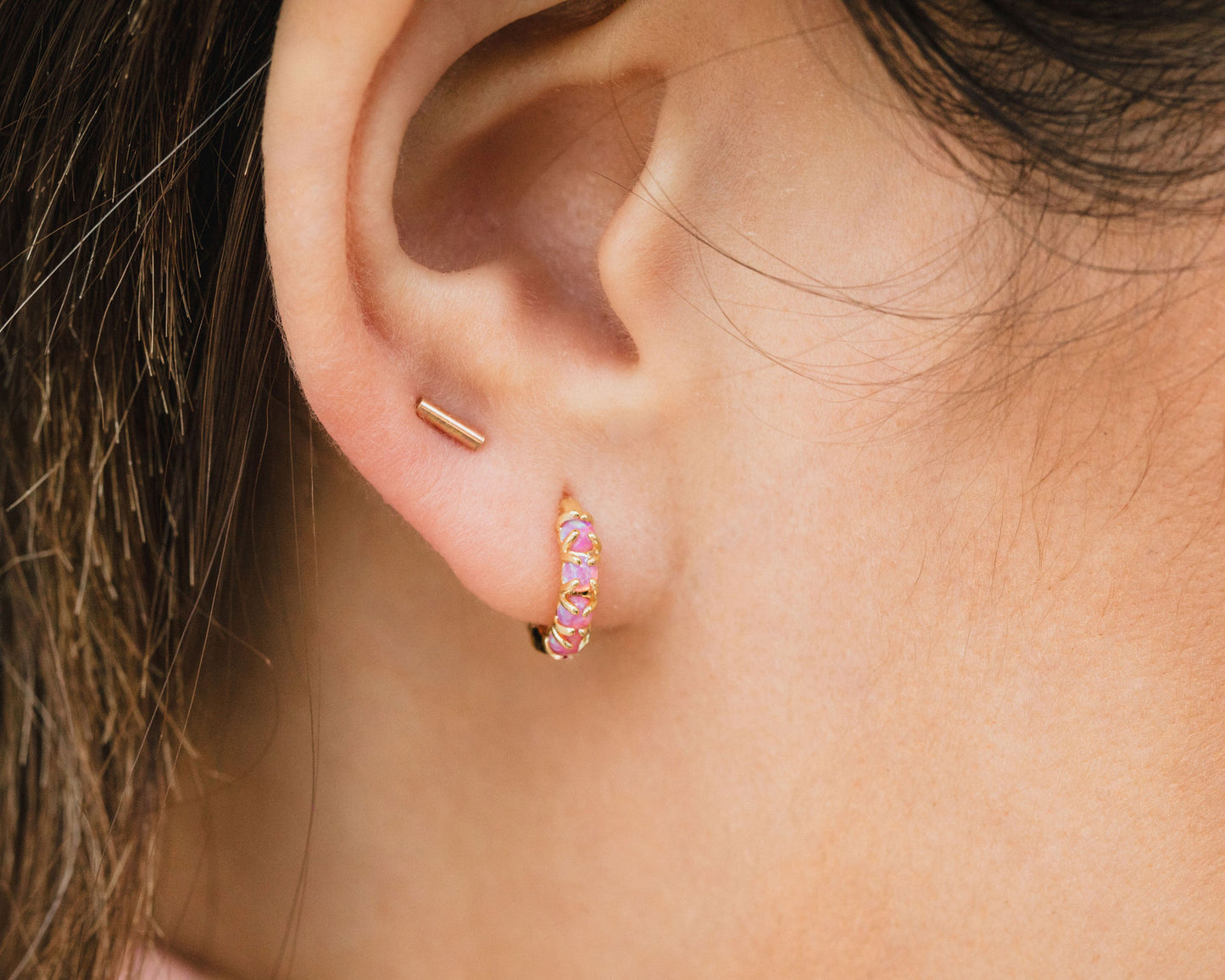 A close-up view of a person's ear adorned with two gold earrings, one of which is a small hoop earring with a pink gemstone.