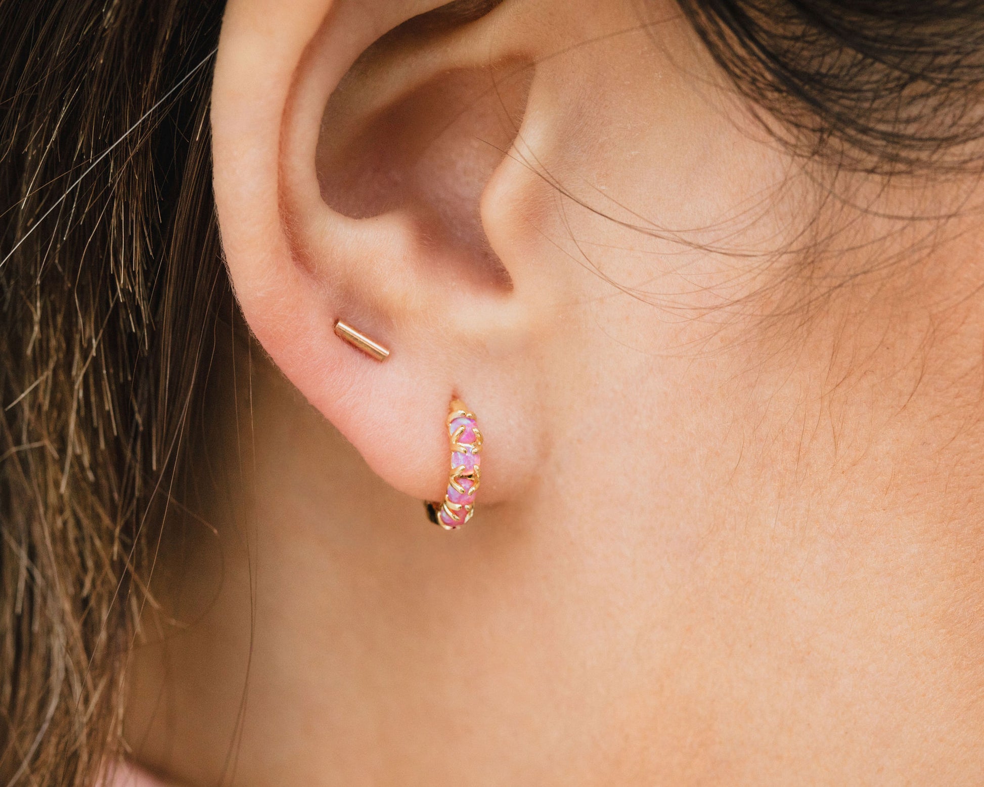 A close-up view of a person's ear adorned with two gold earrings, one of which is a small hoop earring with a pink gemstone.