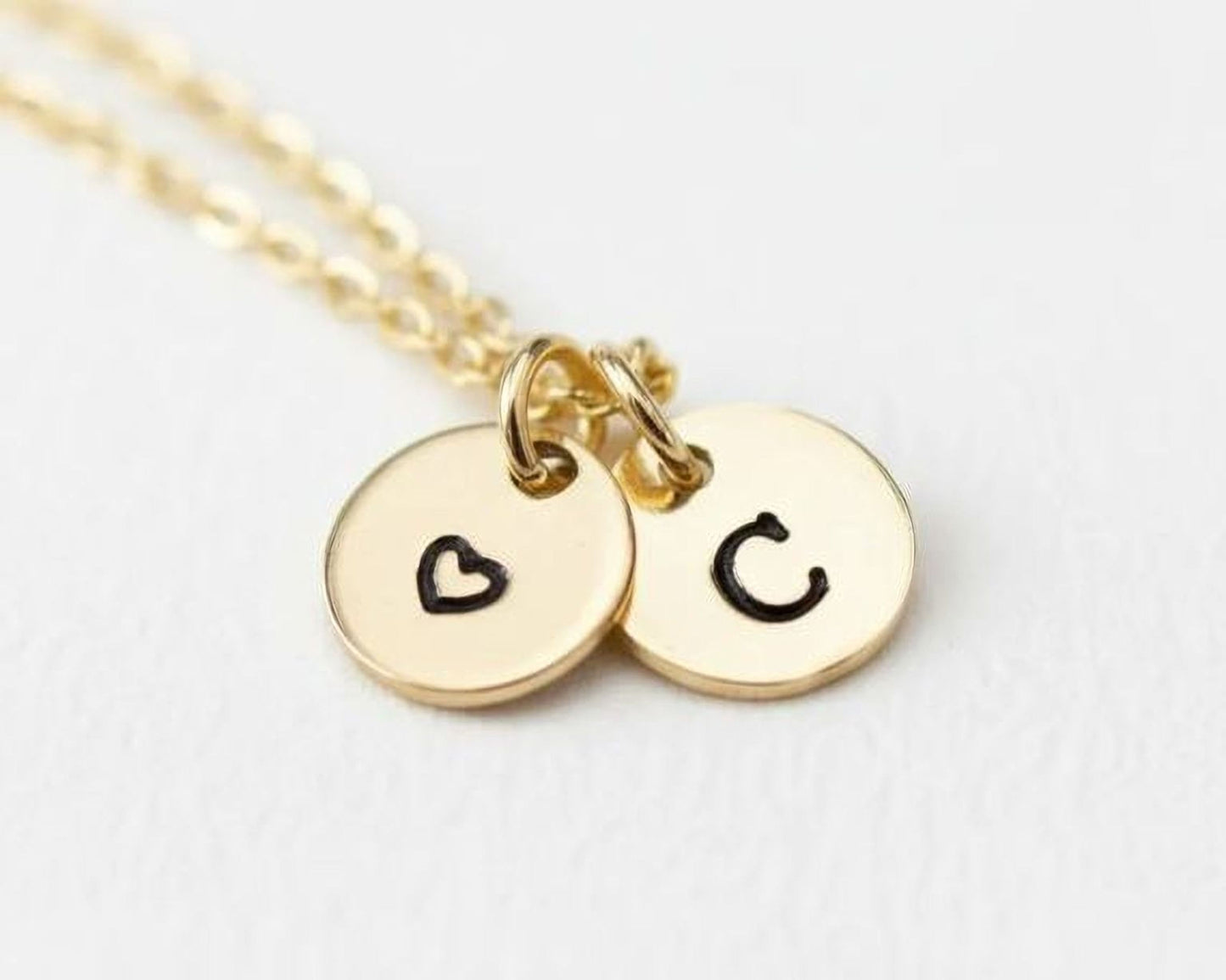 A gold-colored necklace with two round charms attached to it, each with a black heart symbol and the letter 'C' printed on them.