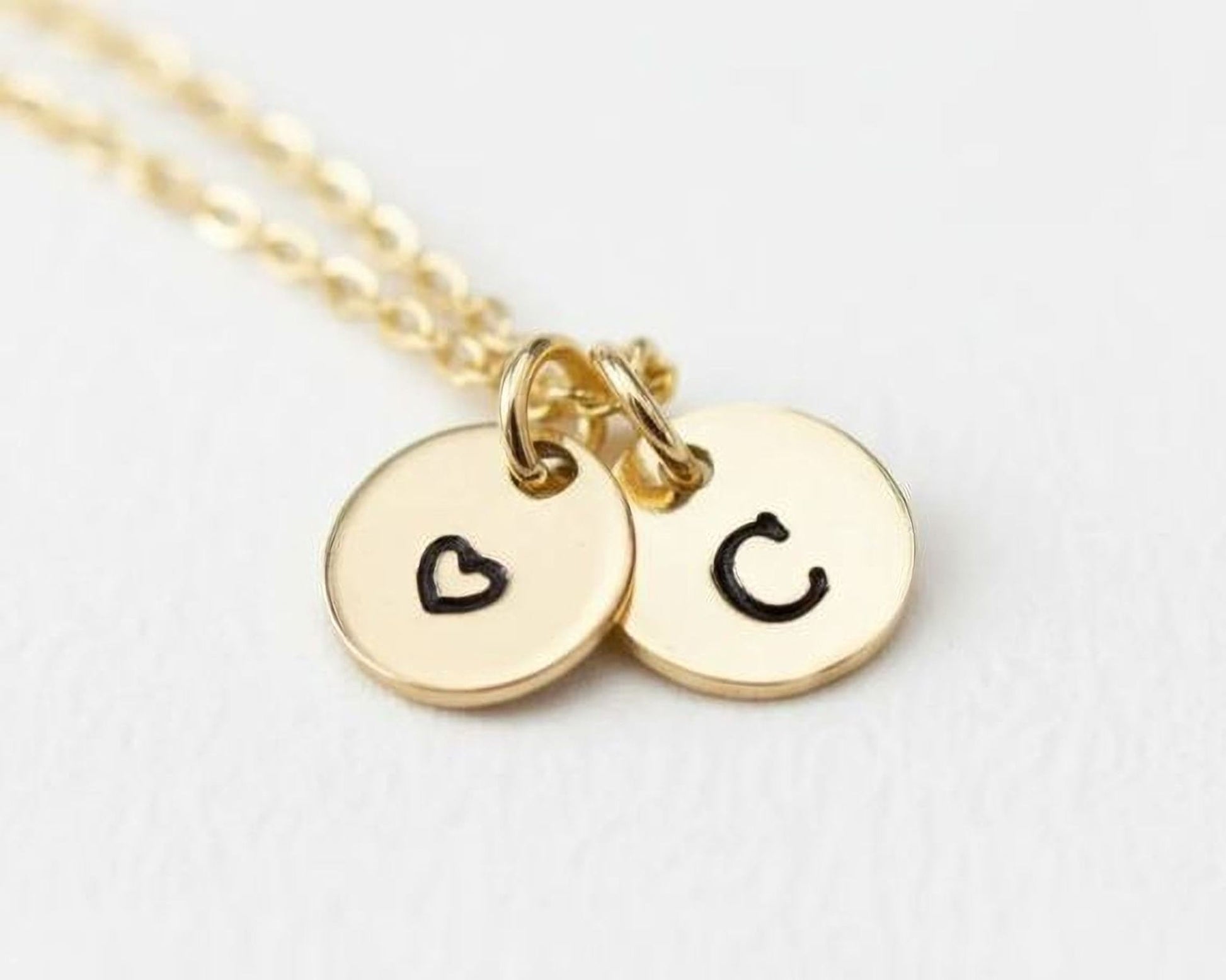 A gold-colored necklace with two round charms attached to it, each with a black heart symbol and the letter 'C' printed on them.
