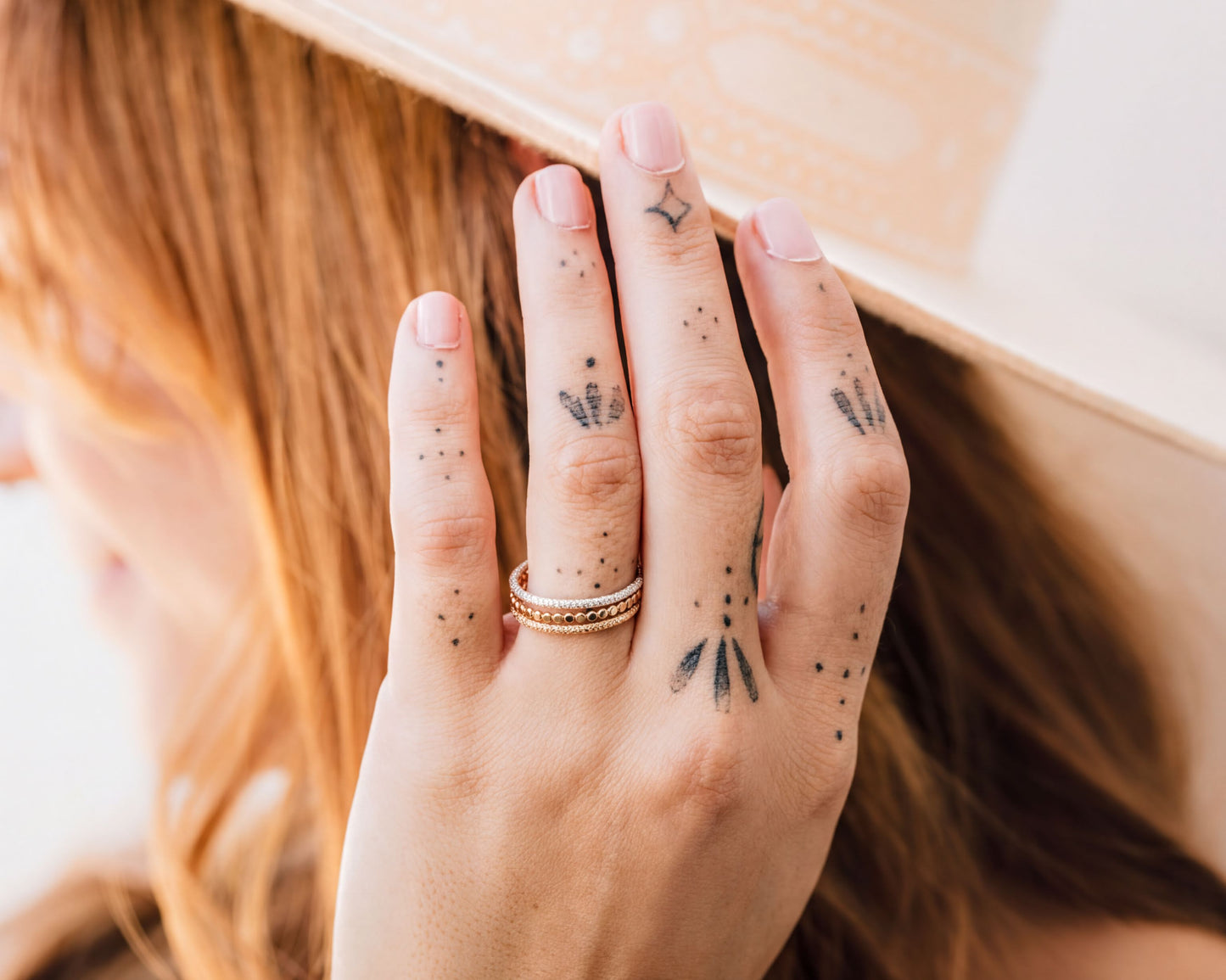 A close-up of a person's hand with tattoos on the fingers, wearing a gold ring, and partially covering their face.