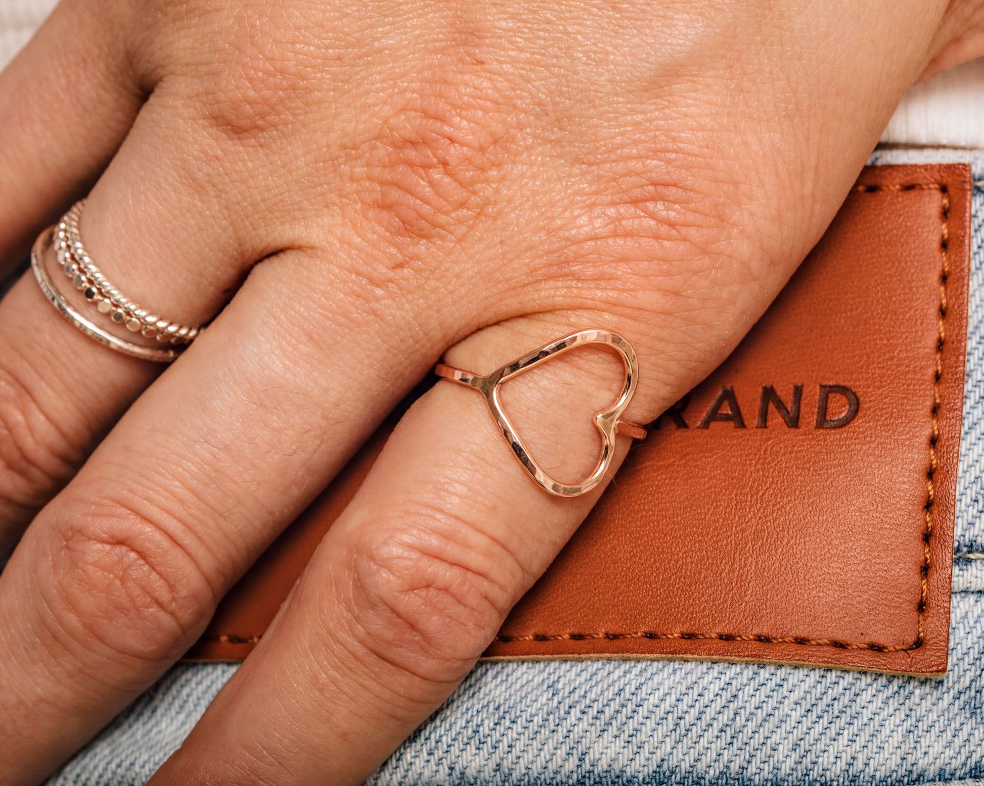 A close-up image of a person's hand wearing a heart-shaped ring, with a leather wallet or card visible in the background.