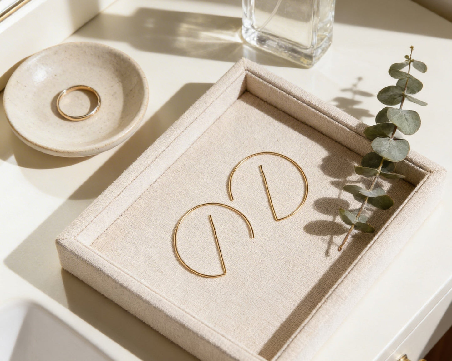 a beige tray with a pair of gold earrings and a ring on it, along with a small white bowl and a clear bottle.