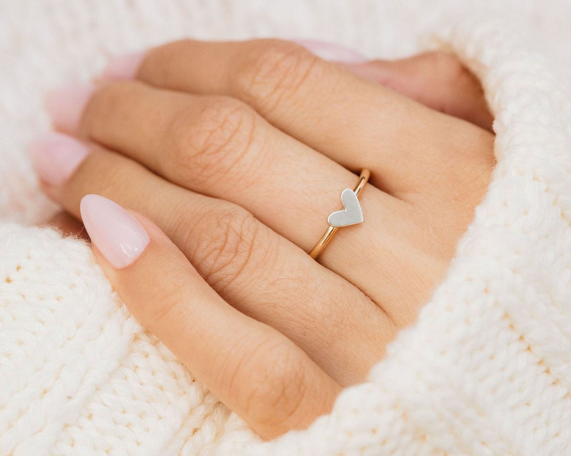 A close-up of a person's hand wearing a gold ring with a heart-shaped accent, resting on a white knitted fabric.