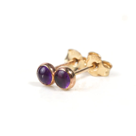 A pair of gold earrings with purple gemstones, specifically amethyst, are shown against a white background.