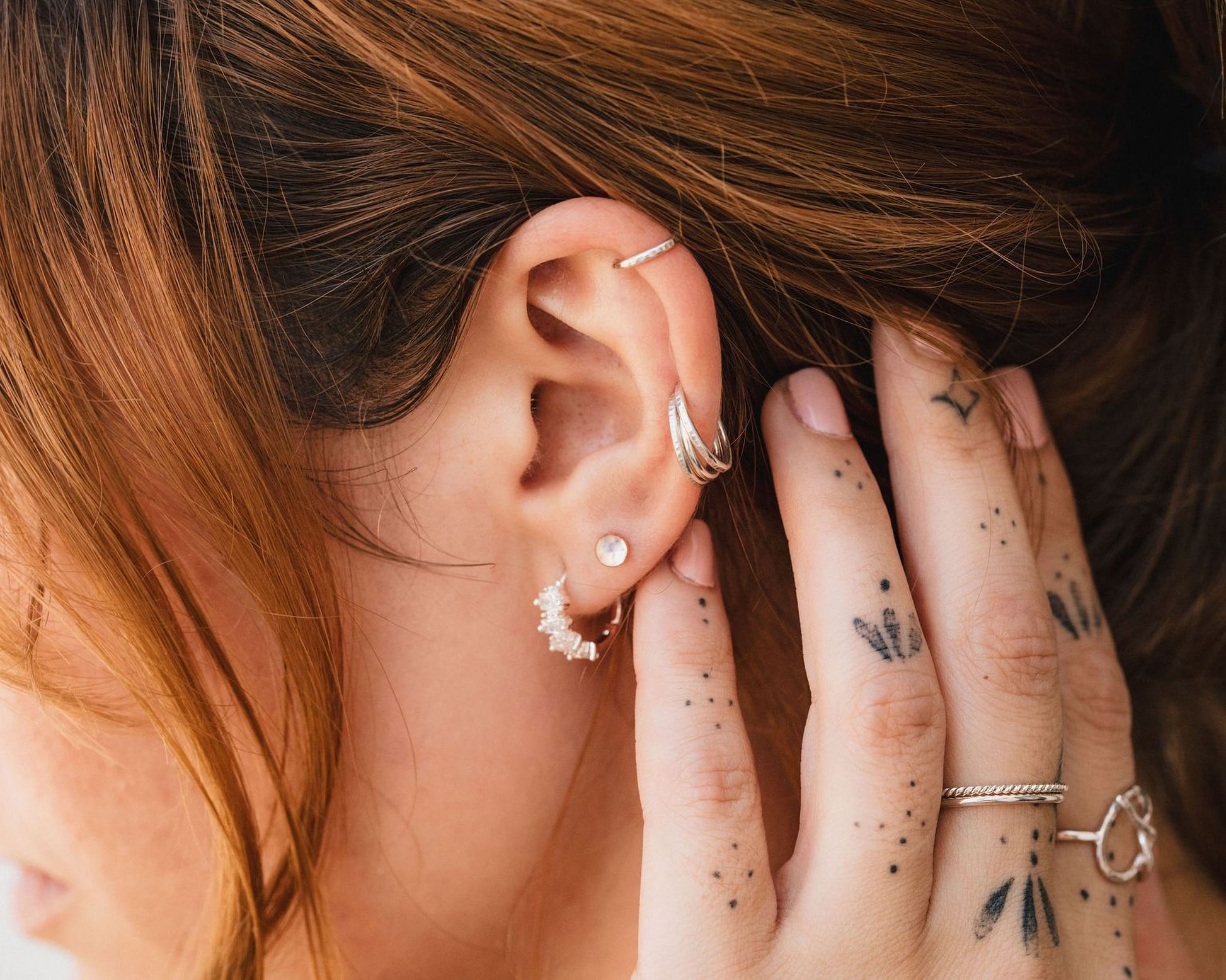 A close-up of a person's hand adorned with multiple rings and earrings, with tattoos visible on the fingers.