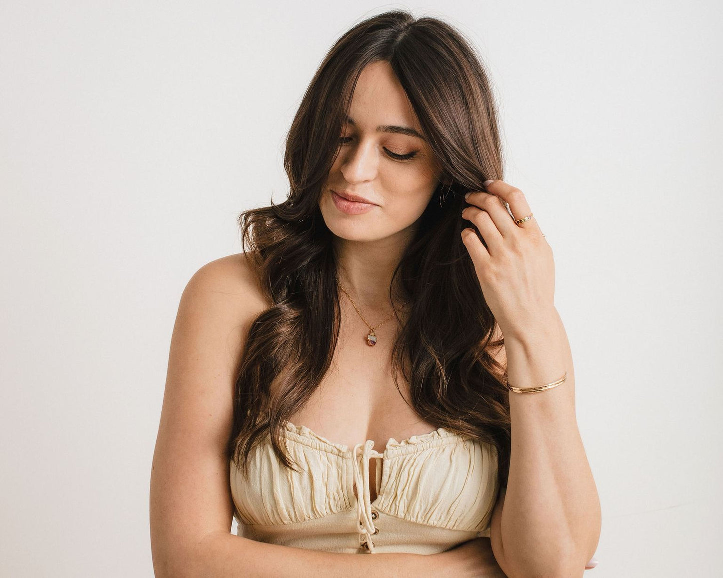 A woman with long brown hair is posing for a portrait, looking down at her hands. She is wearing a beige strapless top and has a necklace around her neck.