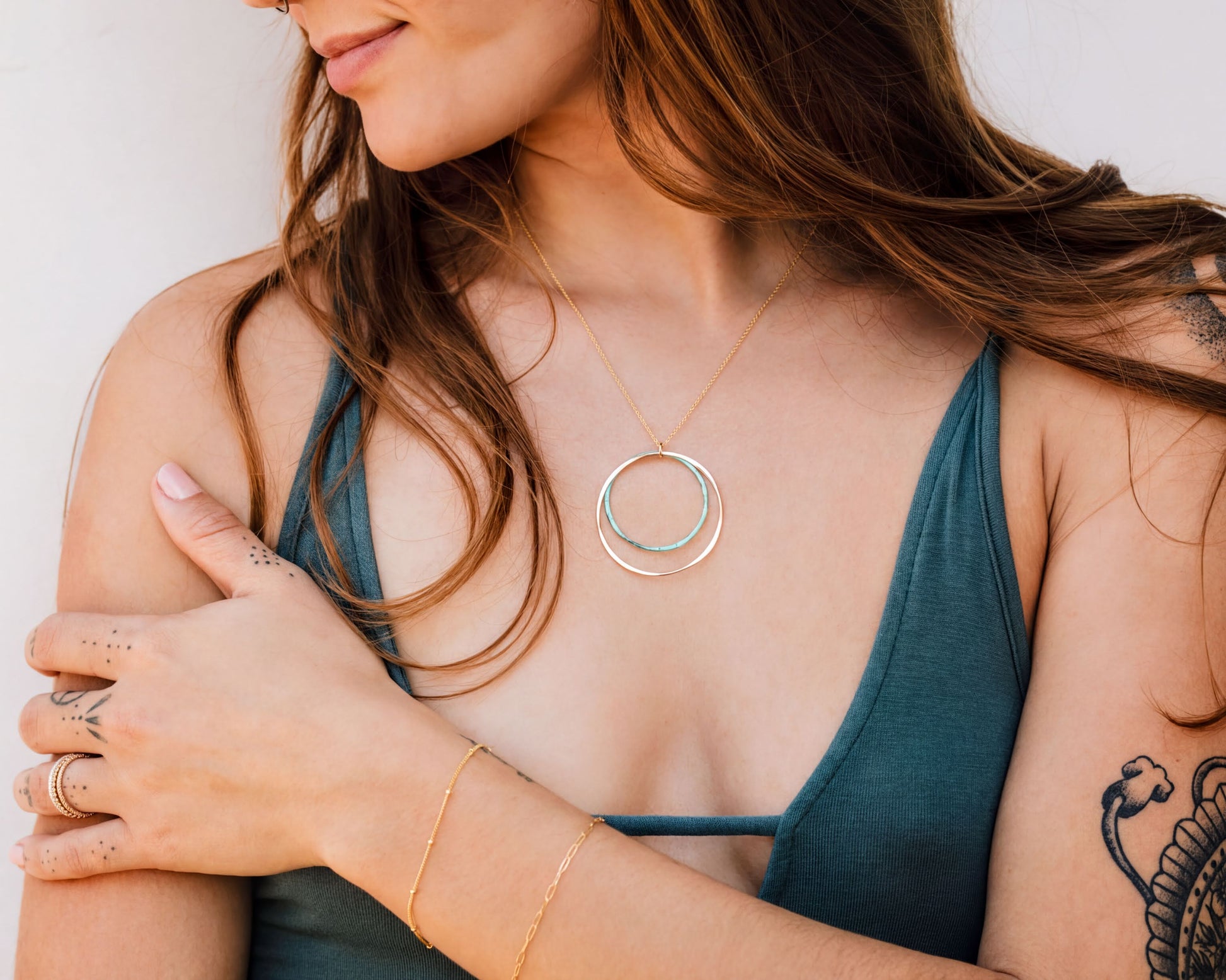 A woman with long brown hair wearing a green top and a necklace with a circular pendant stands with her arms crossed.