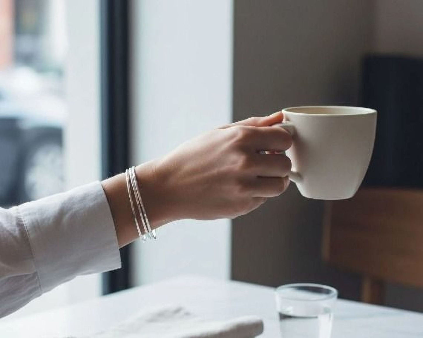 A person's hand holding a white mug, with a glass of water and a napkin on a table in front of them.