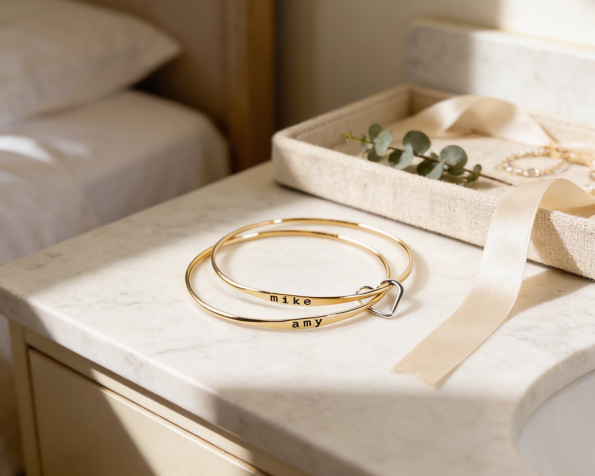 a pair of gold bracelets with the names 'Nike' and 'amy' engraved on them, placed on a white marble countertop next to a small box with a ribbon.