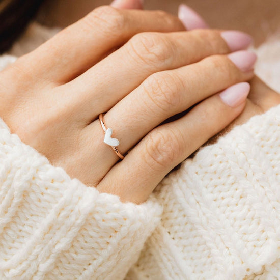 A close-up of a person's hand wearing a gold ring with a heart-shaped accent, resting on a white knitted sweater.