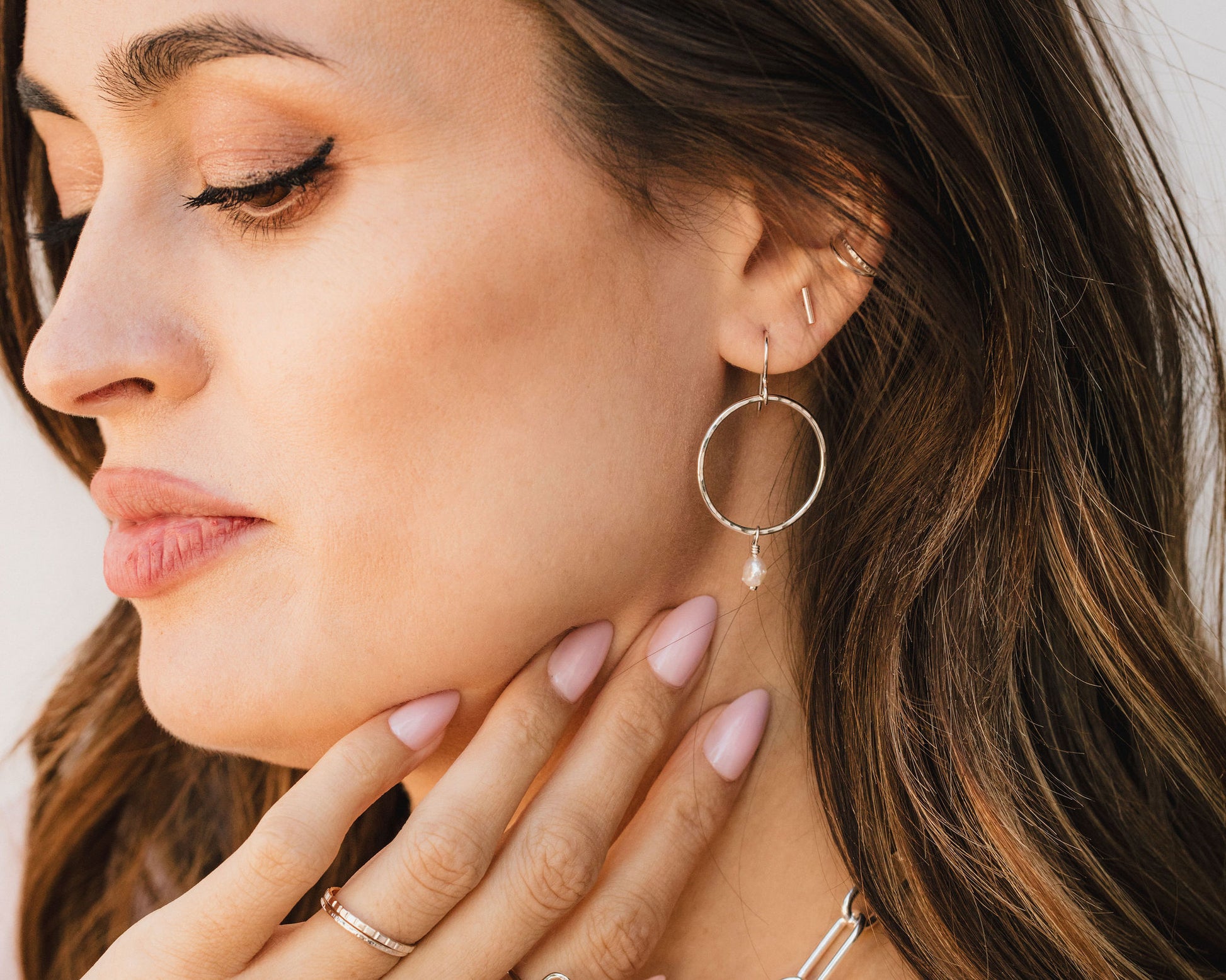 A close-up of a woman's face, with her eyes closed and her hand resting on her chin. She is wearing a pair of hoop earrings and has long, wavy brown hair.