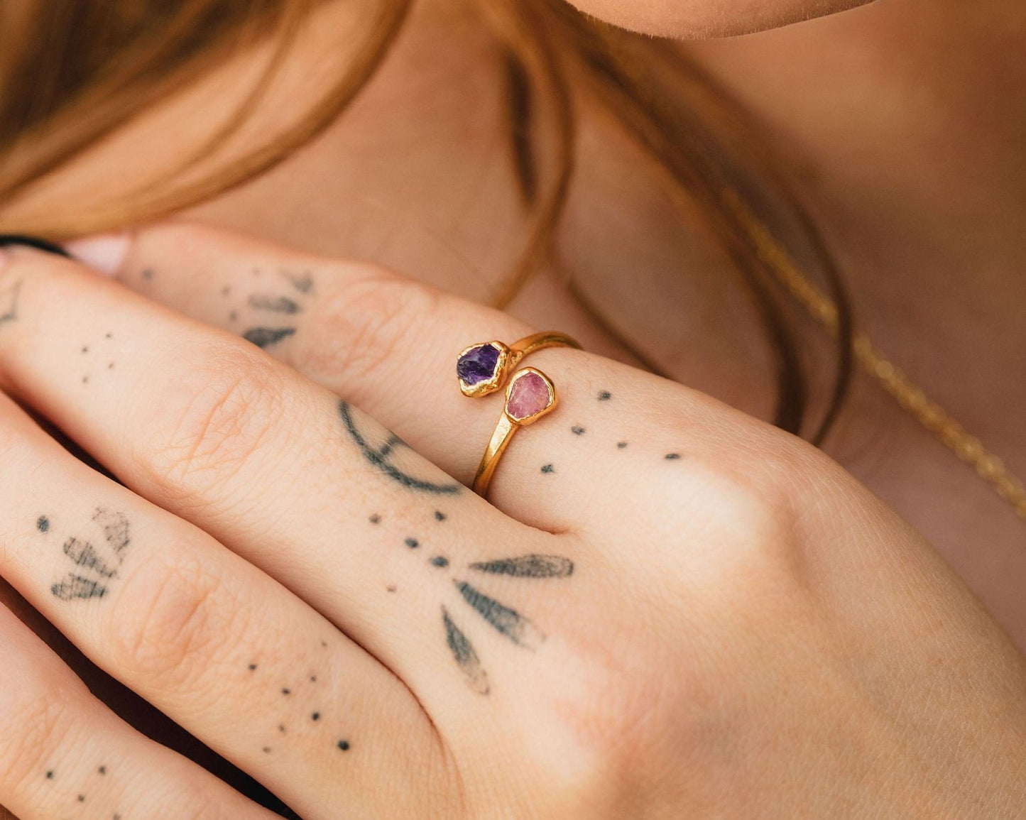 A close-up of a person's hand wearing a gold ring with two gemstones, one purple and one pink, with tattoos on the fingers.