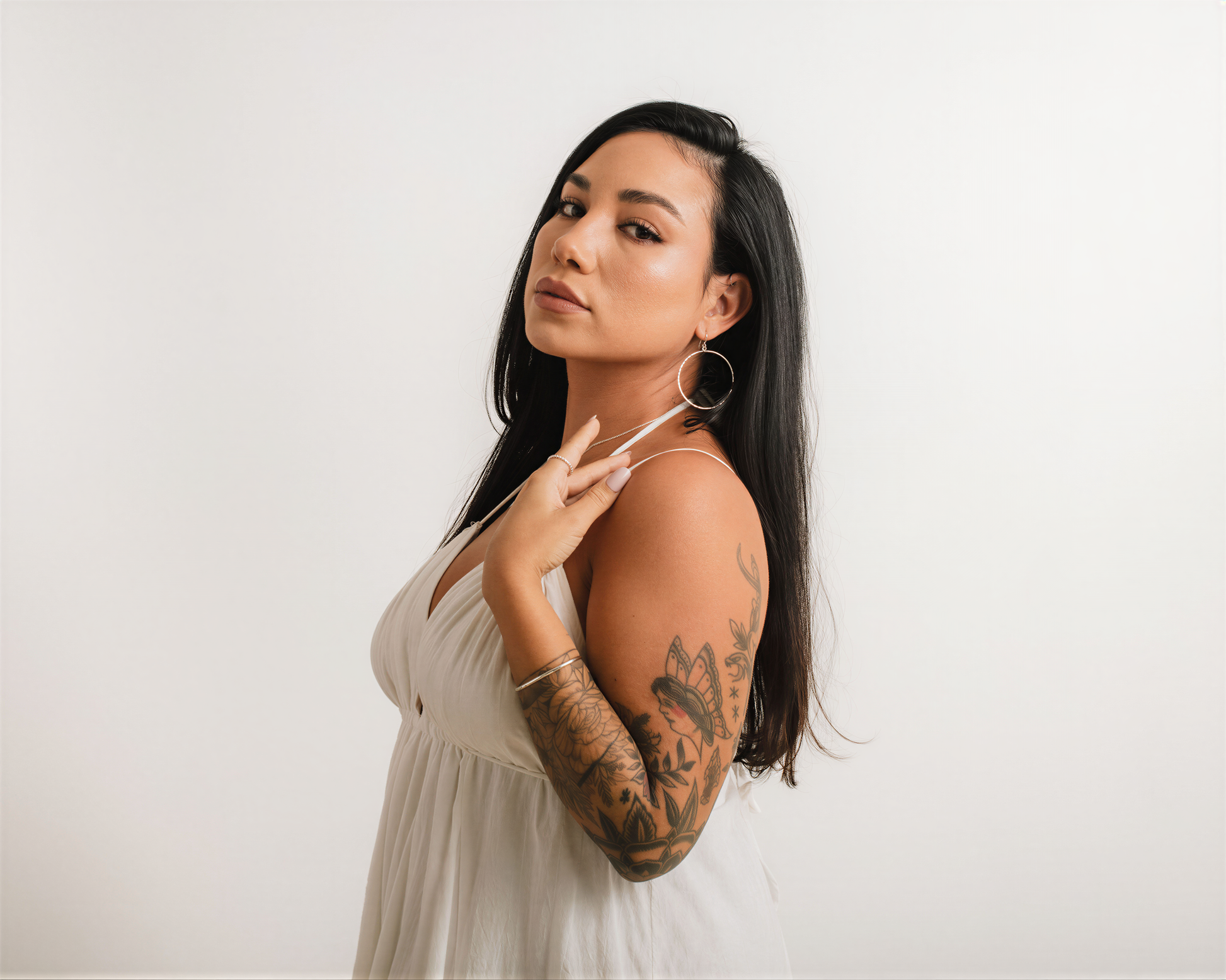 A woman with long dark hair and tattoos poses in a white dress against a plain white background.