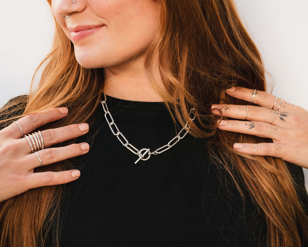 A woman with long, reddish-brown hair wearing a black top and a silver necklace with a chain and a clasp. She is smiling and has her hands on her shoulders.
