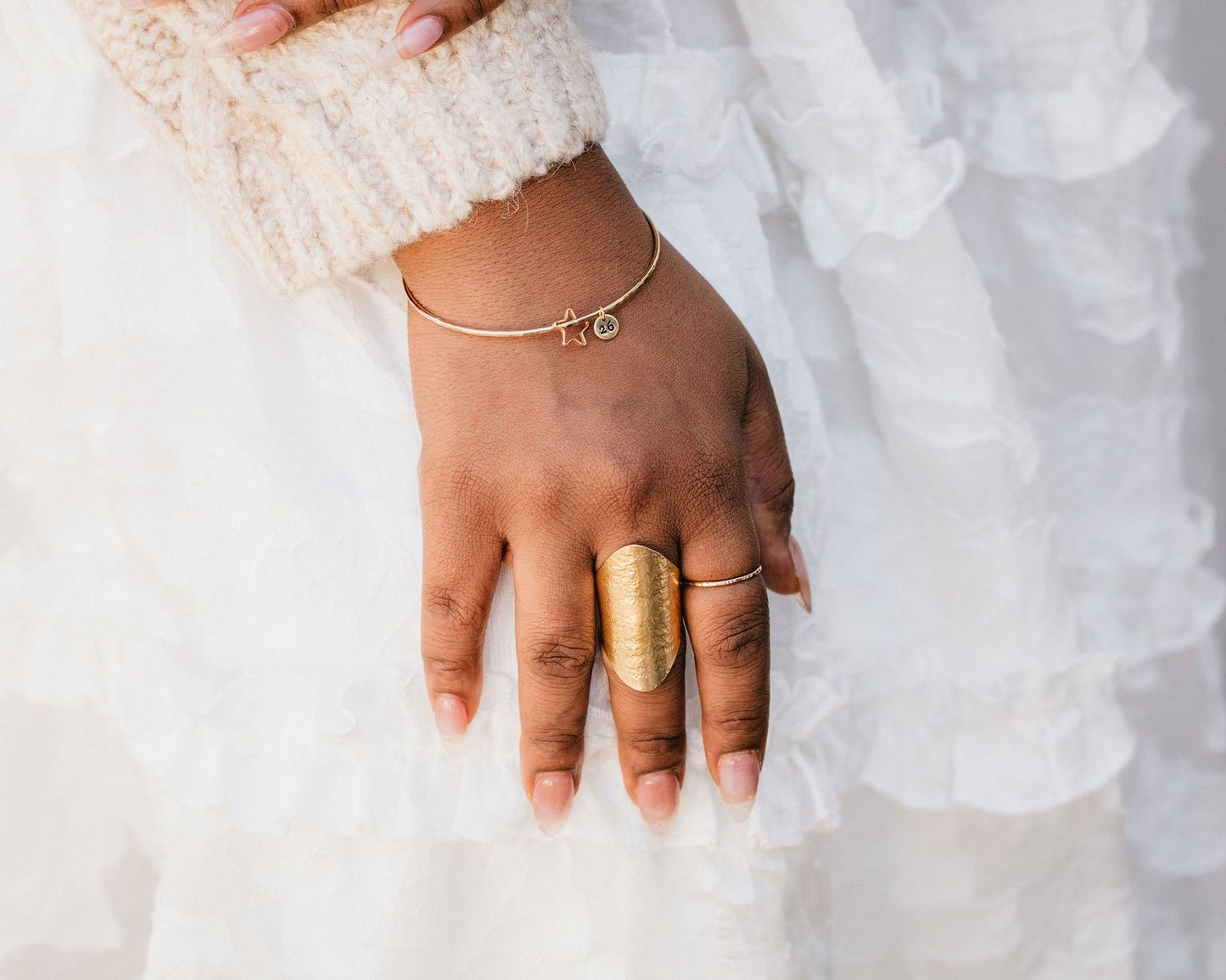 A close-up view of a person's hand wearing a gold ring and bracelet, with a white ruffled skirt visible in the background.