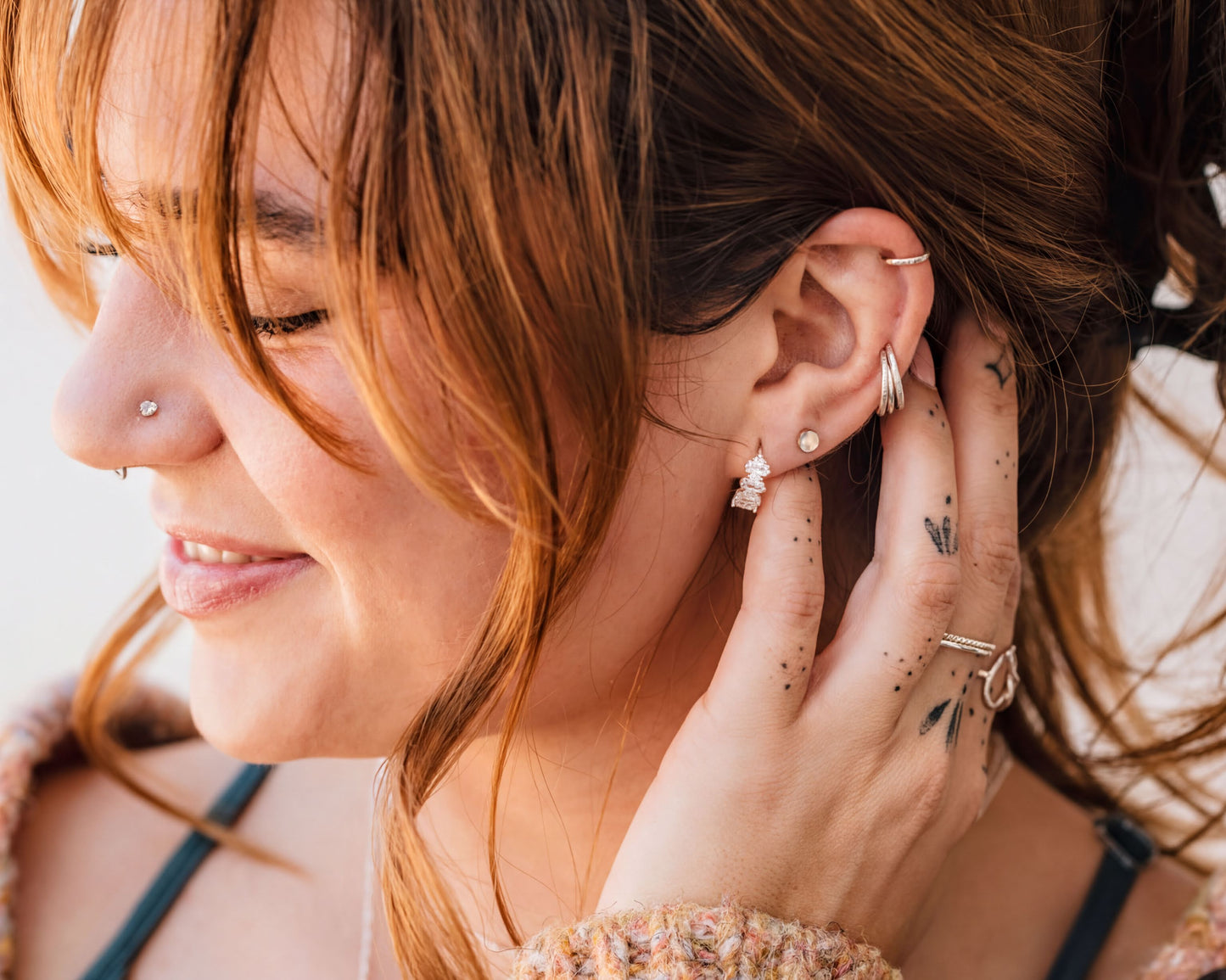 A woman with long brown hair and multiple piercings is smiling and touching her ear with her hand.