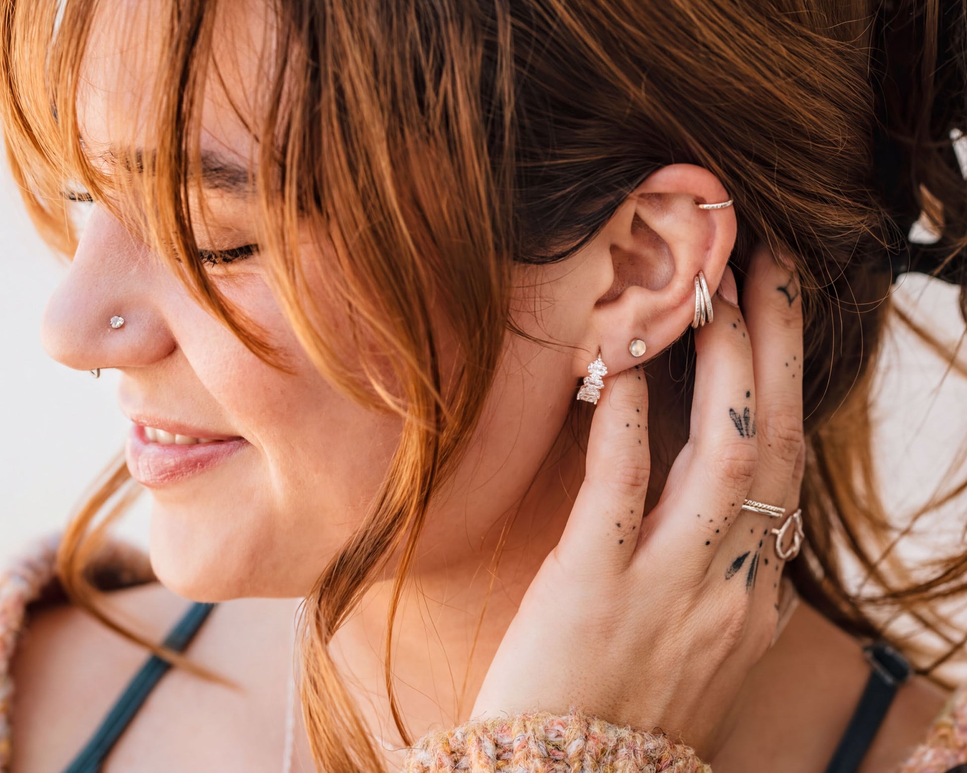 A woman with long brown hair and multiple piercings is smiling and touching her ear with her hand.