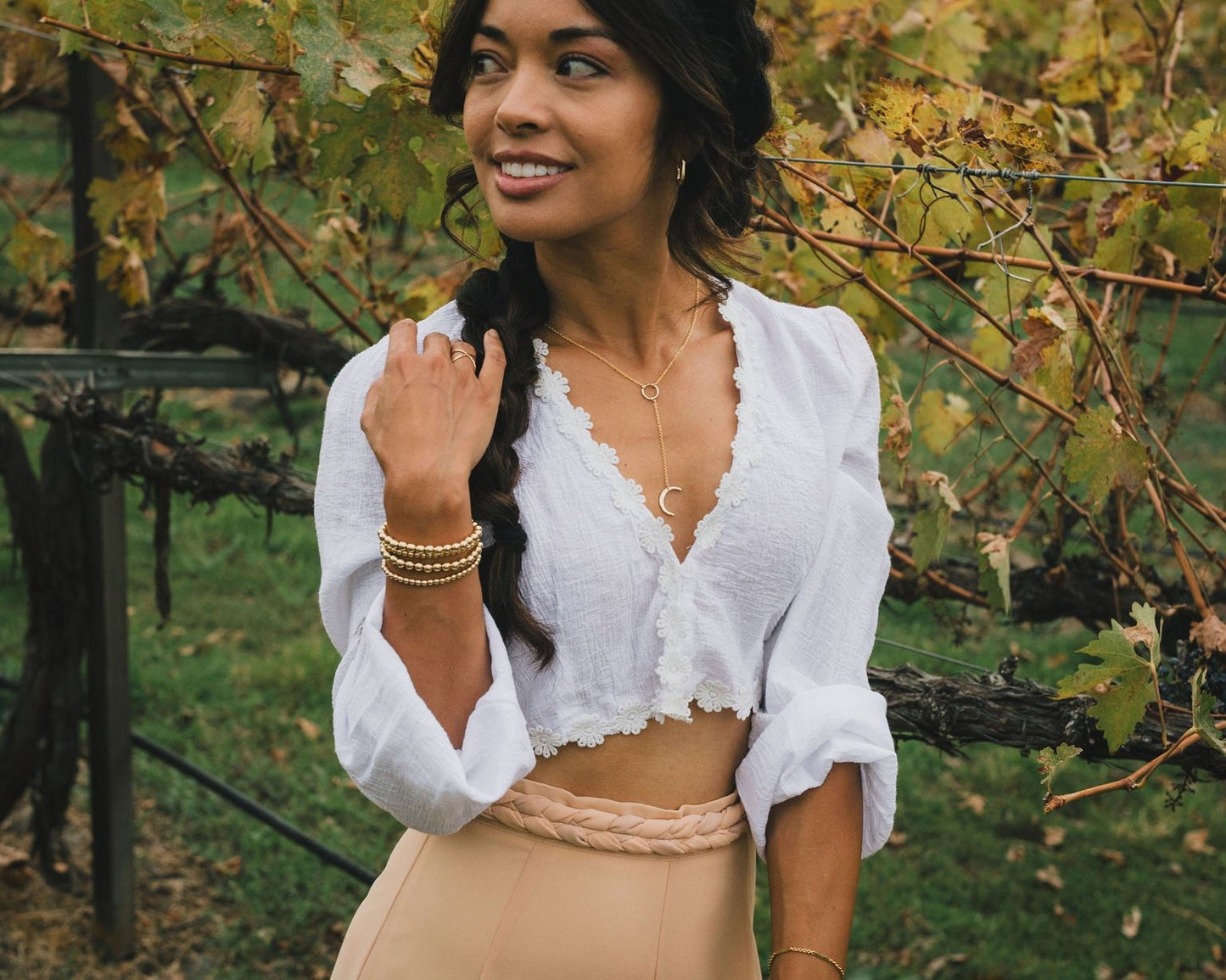 A woman with dark hair is standing in a vineyard, smiling and looking to her left. She is wearing a white blouse with lace trim and a beige skirt.