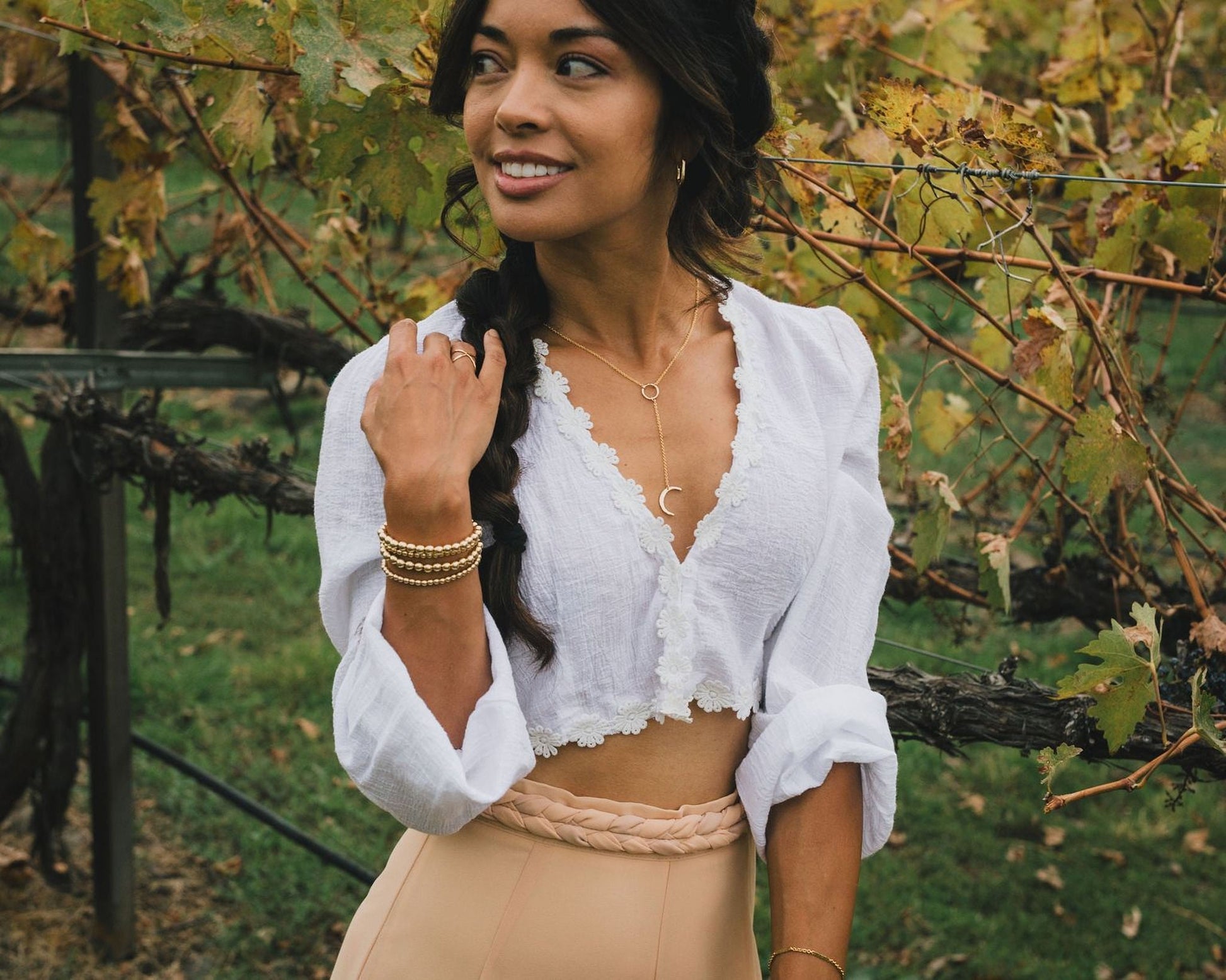 A woman with dark hair is standing in a vineyard, smiling and looking to her left. She is wearing a white blouse with lace trim and a beige skirt.