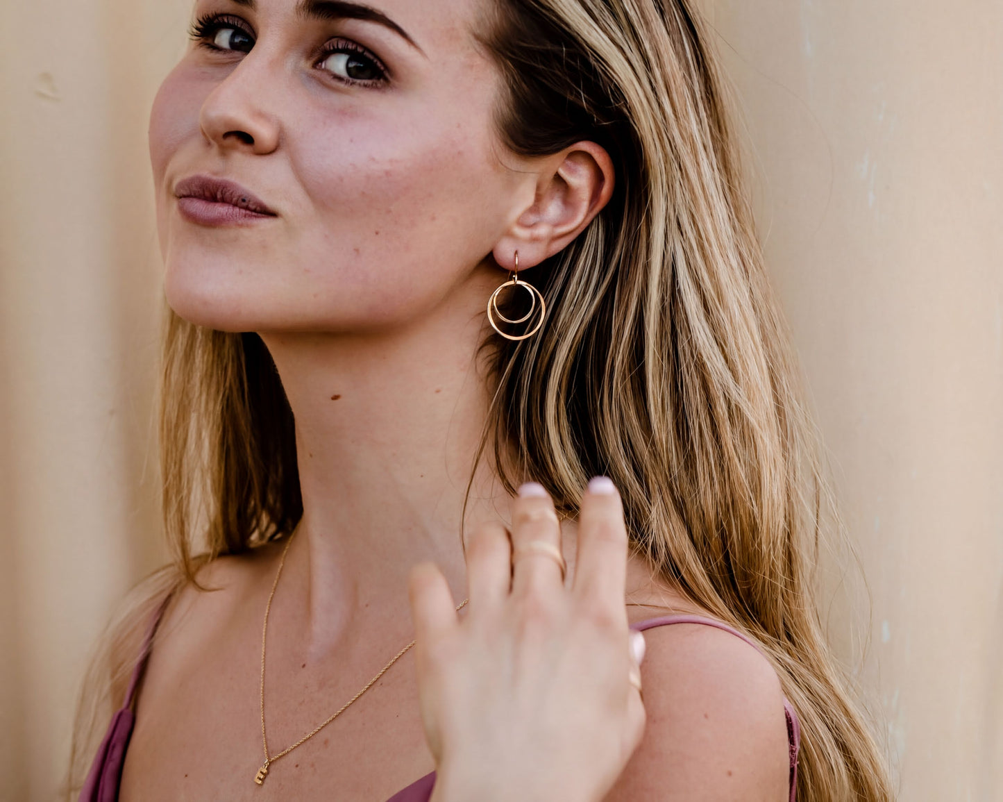 A woman with long blonde hair wearing a gold necklace and earrings, smiling and looking to the side.